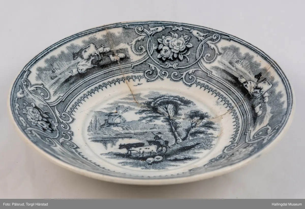 a) Kopp: Stentøy, hvit, liten. Markert fotring. Siden svunget. Utv. polygongal, trykte sorte dekorasjoner. I bunnen innv. landskap. langs randen innv. og utv. 2 blomsterornamenter og 2 felt med kuer og sauer i et slettelandskap. Sprukket.
b) Skål: Stentøy, hvt, rund. Forholdsvis dyp med skrå side. Siden samme dekor som koppen. I bunnen landskap med trær, vindmølle, rytter og kyr. Sprukket og limt. R.B.S.