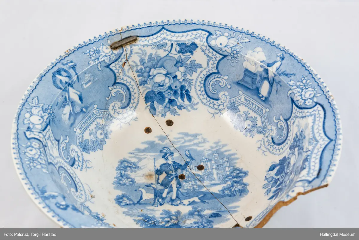 Flint? Hvitt, rundt, fotring. Utbuket side. markert kant med blå dekorasjoner. I bunnen landskap med jeger, symboliserende "april". Veggen innv. og utv. - blomsterklaser omgitt av knekkete bånd, mellom dem kvinnefigurer. Sprukket og klinket. Et stykke mangler. Bunnen stemplet: "Patterson" 12.

R.B.S.