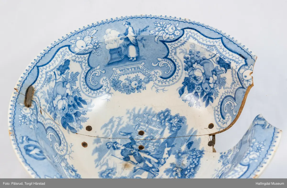 Flint? Hvitt, rundt, fotring. Utbuket side. markert kant med blå dekorasjoner. I bunnen landskap med jeger, symboliserende "april". Veggen innv. og utv. - blomsterklaser omgitt av knekkete bånd, mellom dem kvinnefigurer. Sprukket og klinket. Et stykke mangler. Bunnen stemplet: "Patterson" 12.

R.B.S.