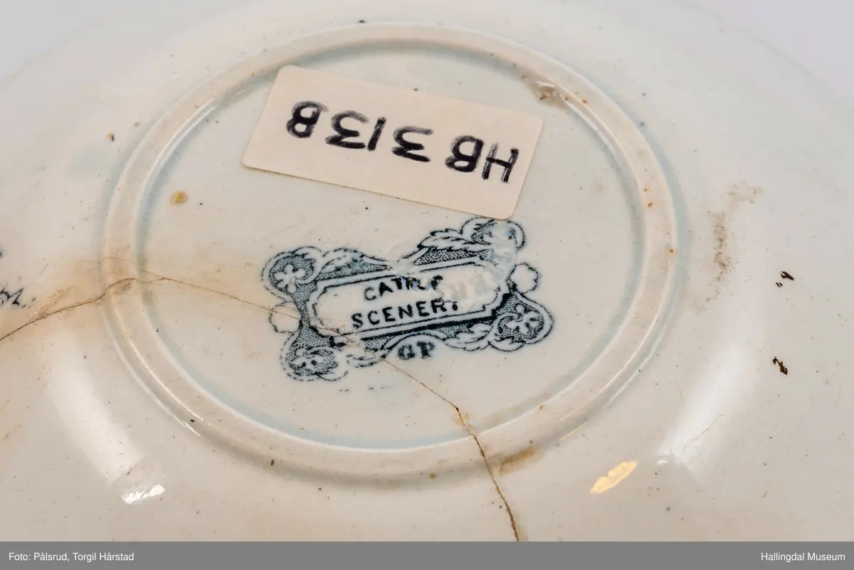 a) Kopp: Stentøy, hvit, liten. Markert fotring. Siden svunget. Utv. polygongal, trykte sorte dekorasjoner. I bunnen innv. landskap. langs randen innv. og utv. 2 blomsterornamenter og 2 felt med kuer og sauer i et slettelandskap. Sprukket.
b) Skål: Stentøy, hvt, rund. Forholdsvis dyp med skrå side. Siden samme dekor som koppen. I bunnen landskap med trær, vindmølle, rytter og kyr. Sprukket og limt. R.B.S.