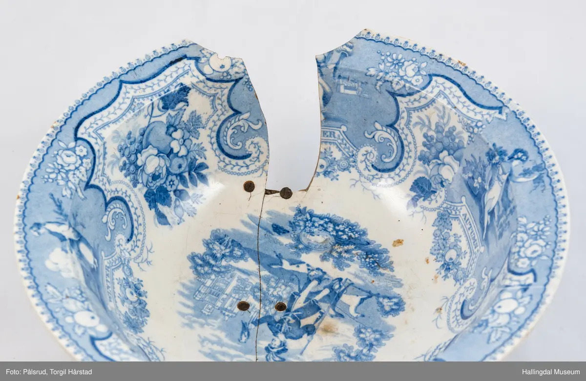 Flint? Hvitt, rundt, fotring. Utbuket side. markert kant med blå dekorasjoner. I bunnen landskap med jeger, symboliserende "april". Veggen innv. og utv. - blomsterklaser omgitt av knekkete bånd, mellom dem kvinnefigurer. Sprukket og klinket. Et stykke mangler. Bunnen stemplet: "Patterson" 12.

R.B.S.