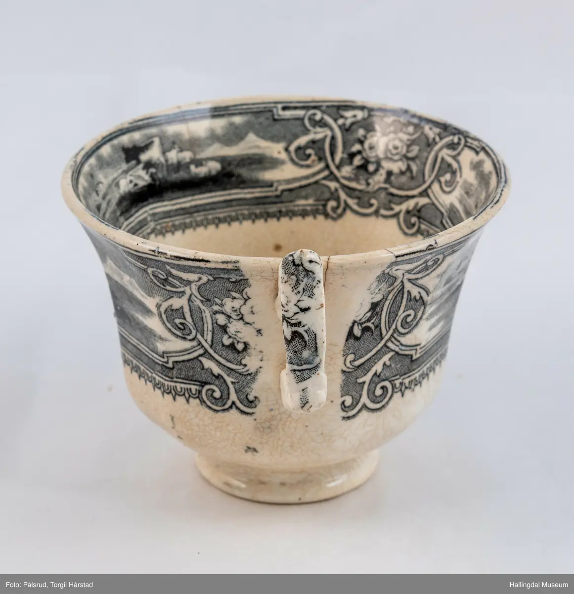 a) Kopp: Stentøy, hvit, liten. Markert fotring. Siden svunget. Utv. polygongal, trykte sorte dekorasjoner. I bunnen innv. landskap. langs randen innv. og utv. 2 blomsterornamenter og 2 felt med kuer og sauer i et slettelandskap. Sprukket.
b) Skål: Stentøy, hvt, rund. Forholdsvis dyp med skrå side. Siden samme dekor som koppen. I bunnen landskap med trær, vindmølle, rytter og kyr. Sprukket og limt. R.B.S.