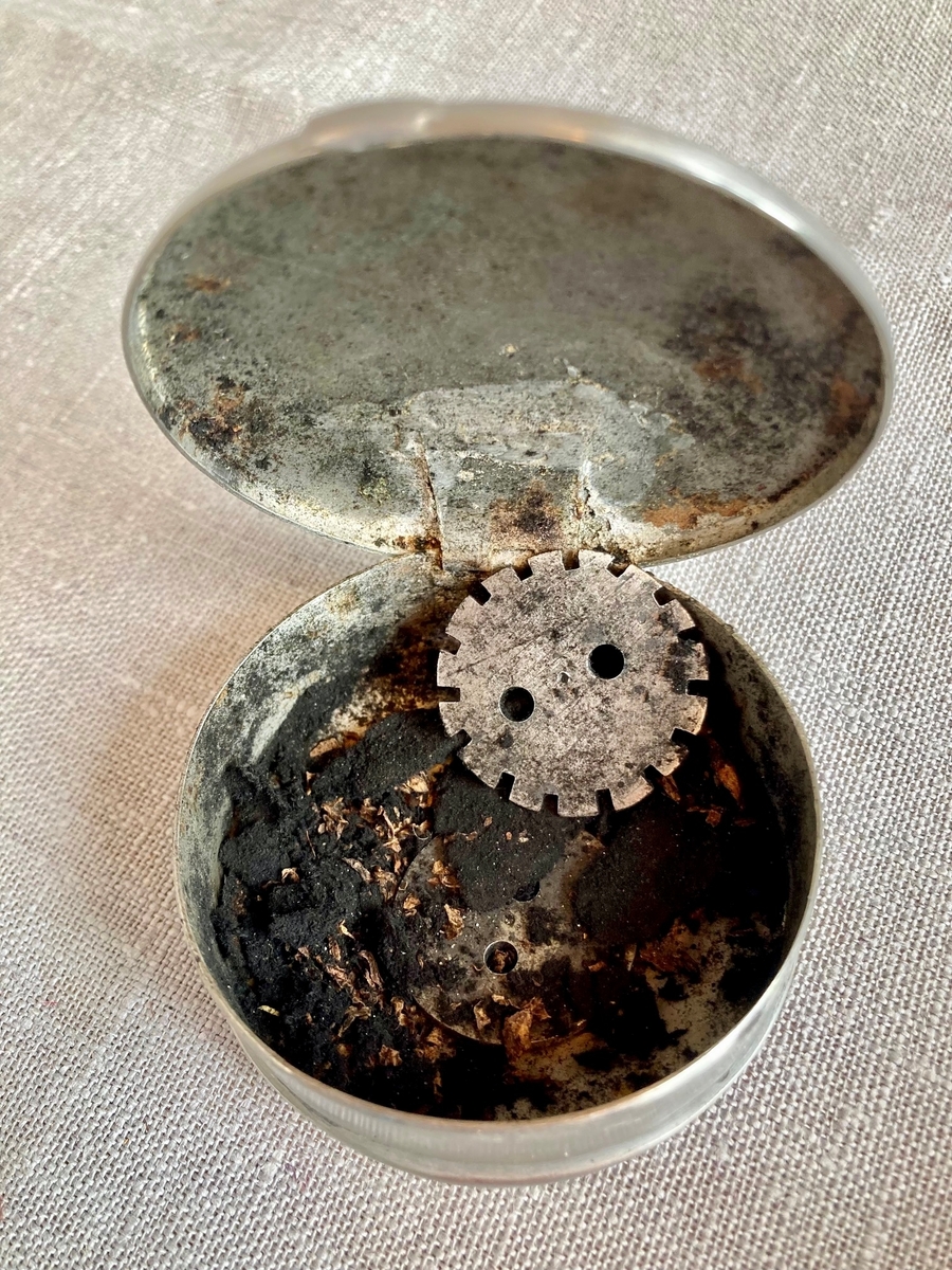 En rund metalleske med hengslet lokk. Ligner en snusdåse. Boksen inneholder noe rusk, trolig tobakk, samt ubestemmelig materiale som ligner rester av sort tekstil, samt små runde rund "knapp" i metall som har tagger rundt kanten.