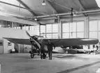 Flygplan M 1, Nieuport IV-G står i hangar på Centrala flygverkstaden Malmslätt. Två män framför flygplanet. I samband med flygdagen på Malmen, 1962.