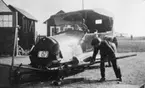 Man i arbete med reparation av en bil tillhörande Flygkompaniet på Malmen, omkring 1920. Registreringsnummer E 1573. I bakgrunden syns motorprovbockar..