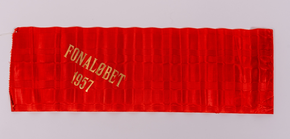 Rött konstsidenband med målad guldtext: "FONALÖBET 1957".