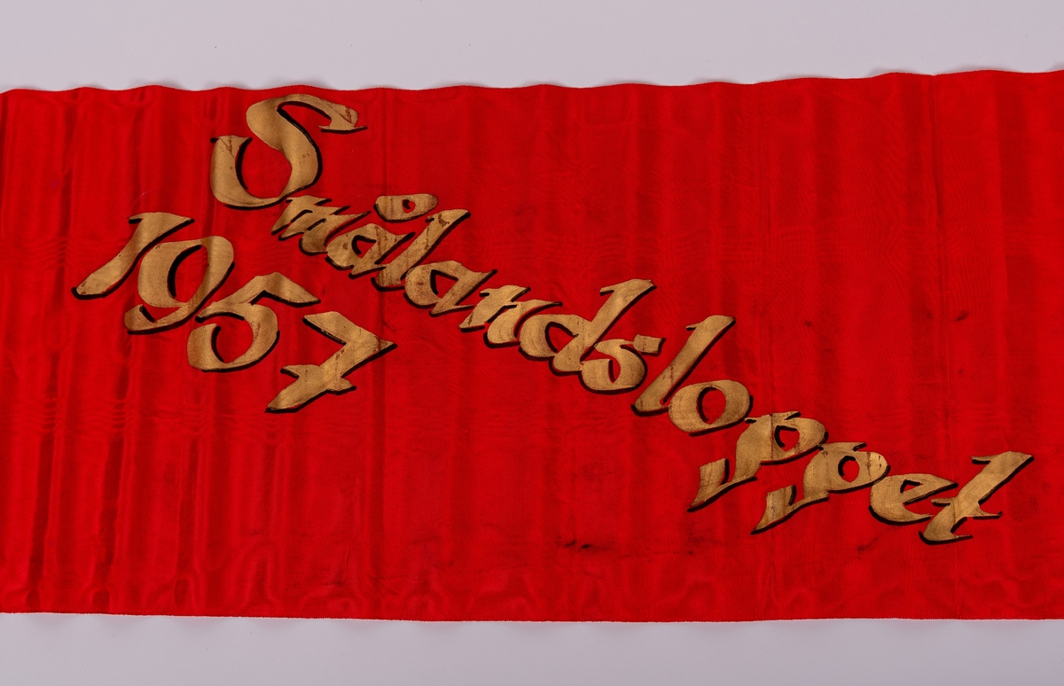 Rött konstsidenband med målad guldtext: "FONALÖBET 1957".