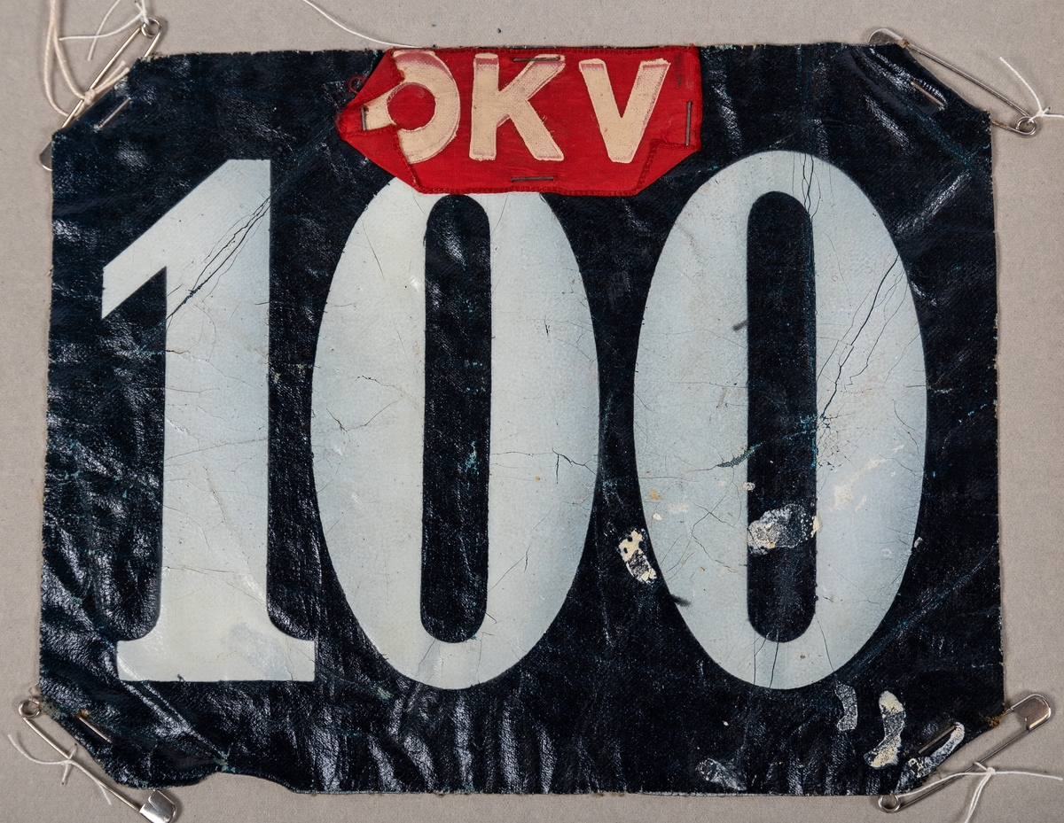 Nummerlappar av galonväv, svart botten med vita siffror: 100. Påsatt bit av rött sidenband med bokstäverna "OKV". Säkerhetsnålar i hörnen för att fästa på kläderna.