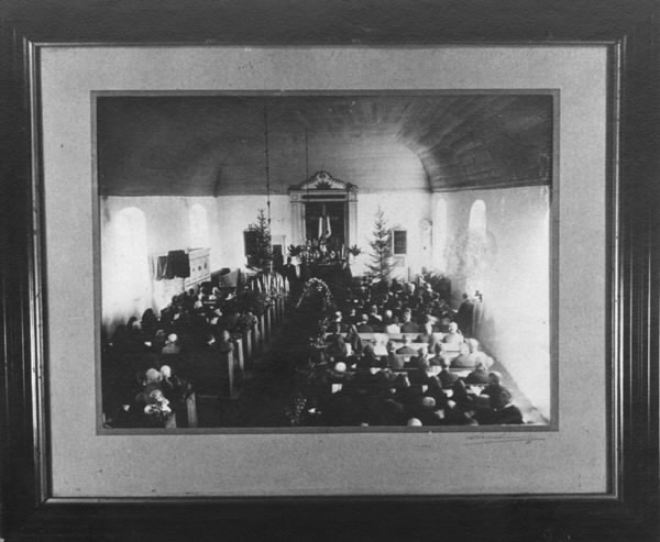 Fotografi inom ram. Föreställande begravning i Drängsereds kyrka. I koret den smyckade kistan. Bänkarna fyllda av människor. Kvinnorna huvudsakligen på vänster sida, männen till höger (syd). Kyrkan prydd med två granar samt björkris. Troligen från 1920-talet. Inramad i en brun träram, guldkant närmast bilden.