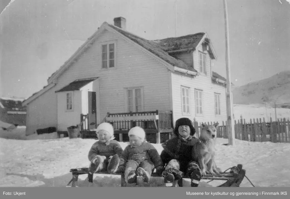 Bildet viser Odny Møllmann, Kari Normann, Ragnhild Normann hunden Sonja som sitter på en kjelke. I bakgrunn ser man familie Normanns bolighus på Melkøya i Finnmark med stakittgjerde og flaggstang. Bildet er trolig tatt i 1937.