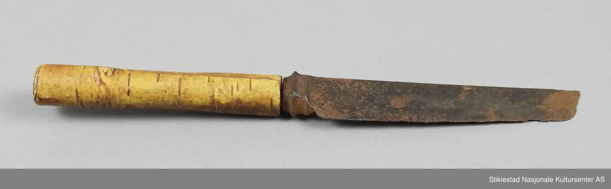 Bordkniv i jern med skaft av bjørkenever. Kniven er antatt fabrikkprodusert. Mange hakk/skader på bladet, en gode del rust. Skaftet er laget av sammenrullet never.