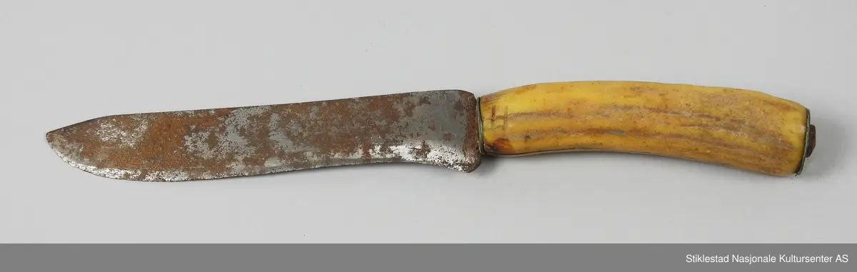 Bordkniv i jern med skaft av reinhorn, antatt heimsmidd? Kniven har en lang tange som går gjennom hele skaftets lengde. I enden av skaftet er det montert en rund plate med hull i midten, tangen går gjennom dette hullet, klinket fast? En god del rust.