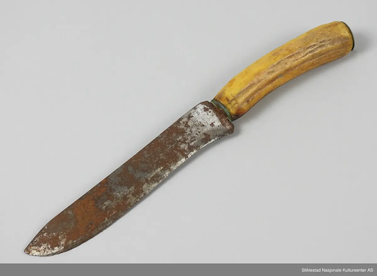 Bordkniv i jern med skaft av reinhorn, antatt heimsmidd? Kniven har en lang tange som går gjennom hele skaftets lengde. I enden av skaftet er det montert en rund plate med hull i midten, tangen går gjennom dette hullet, klinket fast? En god del rust.