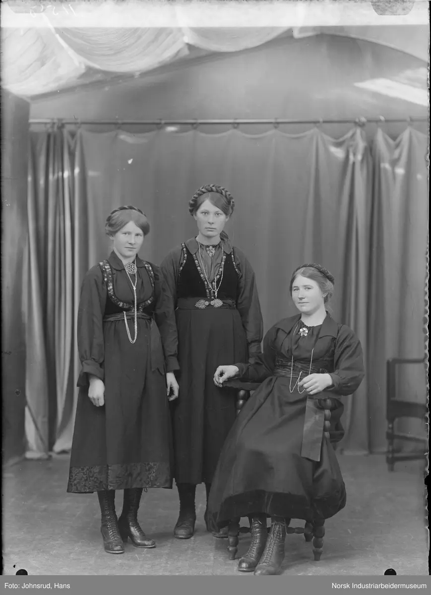Gruppeportrett. Tre kvinner kledd i stakk og liv fotografert i studio. Alle har vippe i håret, smykke rundt halsen og sølje i halsen på skjorta. Anne Finkenhagen sittende til høyre.