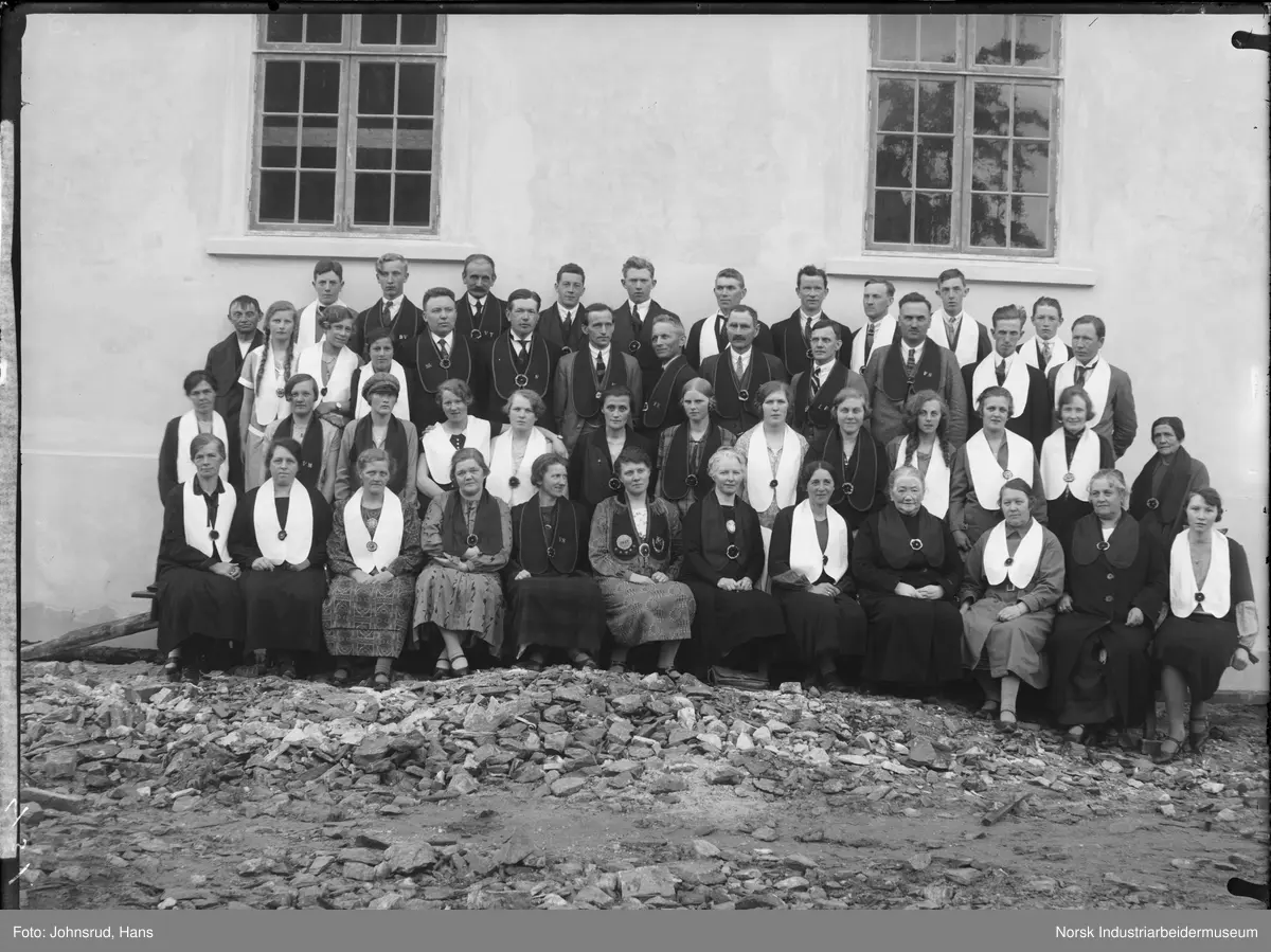 Gruppebilde av IOGT losjen på Rjukan. Kvinner og menn oppstilt foran hus. Alle har ordensbånd over skulderen, noen lyst og noen mørkt, med nål som fester tøystykkene sammen. Kvinnen i midten nederst har logo for IOGT, emblem og stjerner, samt frynser på sitt tøybånd. Flere av de med mørke tøybånd har broderte bokstaver på disse.