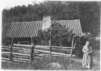 En okänd äldre kvinna med huckle och förkläde står vid en gärdesgård framför ett okänt litet torp.