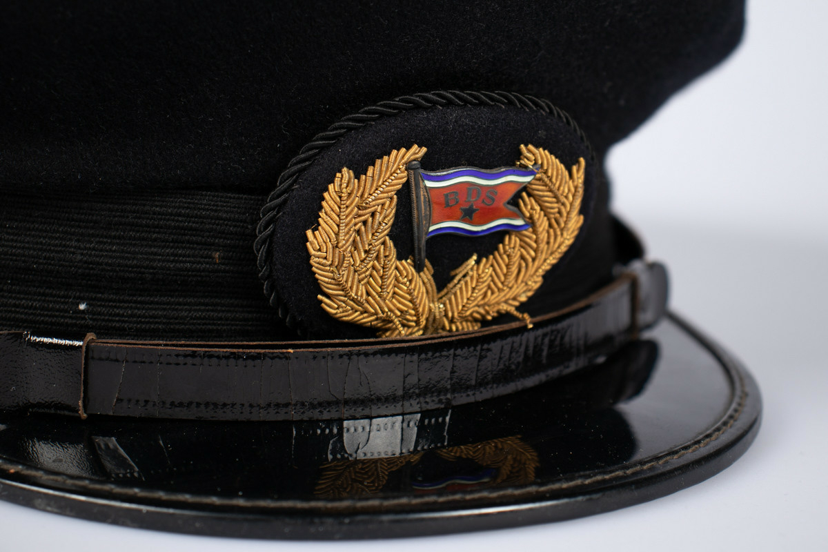 Svart uniformslue med skjerm og rederiflagg på luemerke. To gullfargede knapper i hver side av skjermen  med et svart bånd mellom. Gjennomsiktig plastikk hette er tredd over pullen.