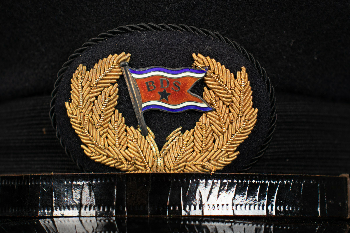 Svart uniformslue med skjerm og rederiflagg på luemerke. To gullfargede knapper i hver side av skjermen  med et svart bånd mellom. Gjennomsiktig plastikk hette er tredd over pullen.