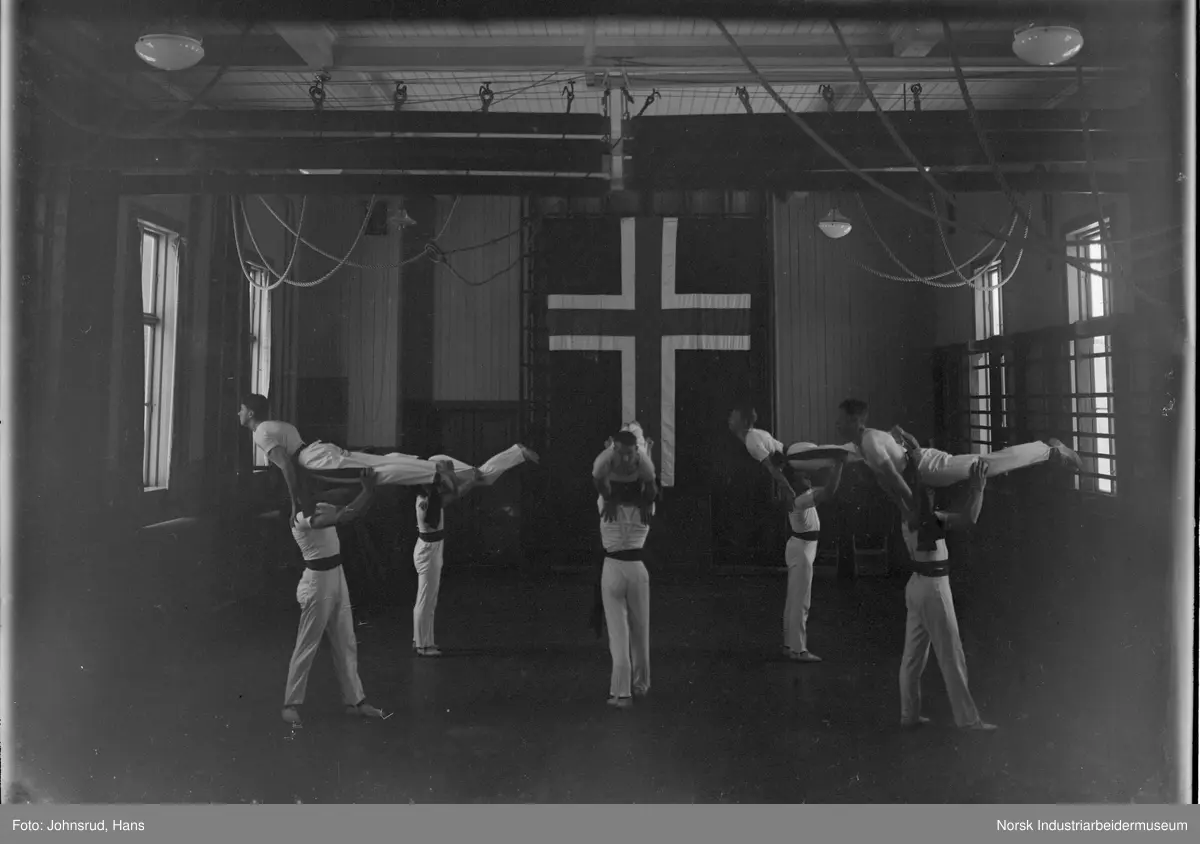 Dobbelportrett av turngruppe fra lærerskolen. 
Venstre bilde: Mannlige turnere i gymsal, fem menn stående på gulvet mens de løfter en mann vannrett over hodet. Norsk flagg på veggen bak i gymsalen. 
Høyre bilde: Ti mannlige turnere oppstilt i pyramideformasjon i gymsal. Ribbevegg og norsk flagg i bakgrunnen.