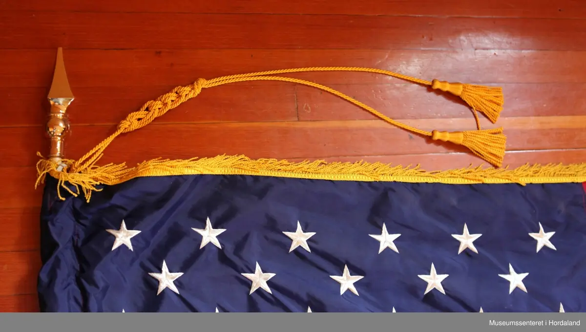 Flagg med stang i to deler, samt en fot. Flagget er rektangulært med 13 røde og hvite horisontale striper, og 50 hvite stjerner på blå bakgrunn i øvre venstre hjørne. Rundt flagget er en gul frynsekant. Flagget er festet til en paradestang av tre med gyllen kule og spyd på toppen. På toppen henger også  en gyllen snor med to dusker som er flettet sammen. Selve stangen kan skrus fra hverandre i to deler.