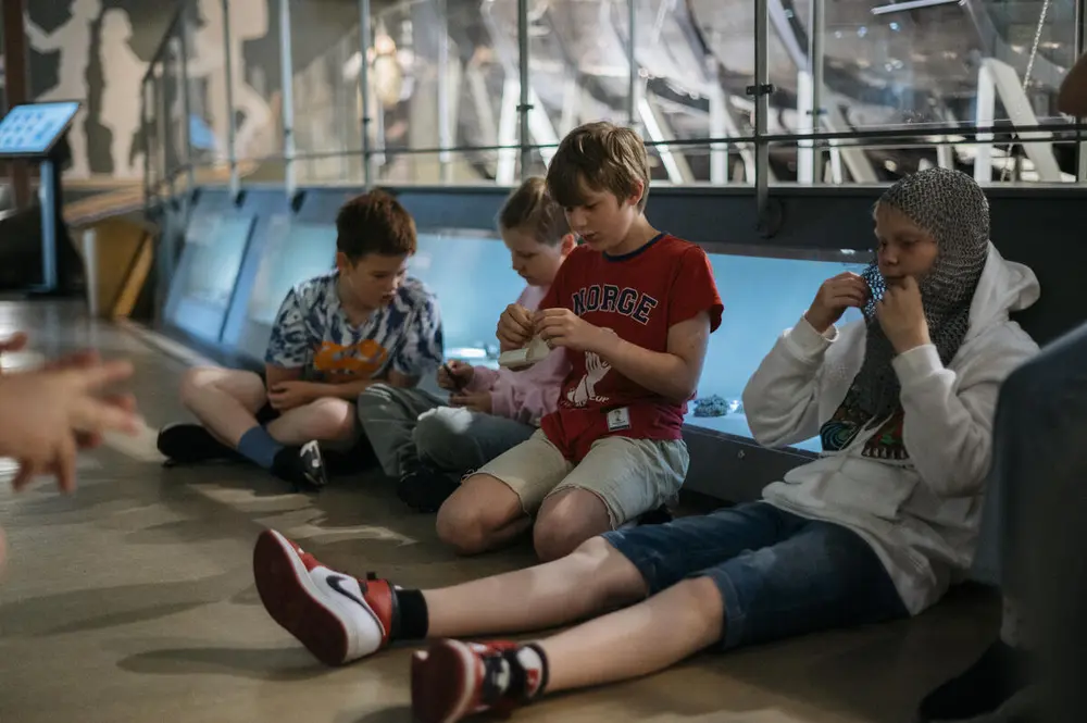 Barn sitter på gulvet i en museumsutstilling. En gutt har på seg et hodeplagg for rustning.