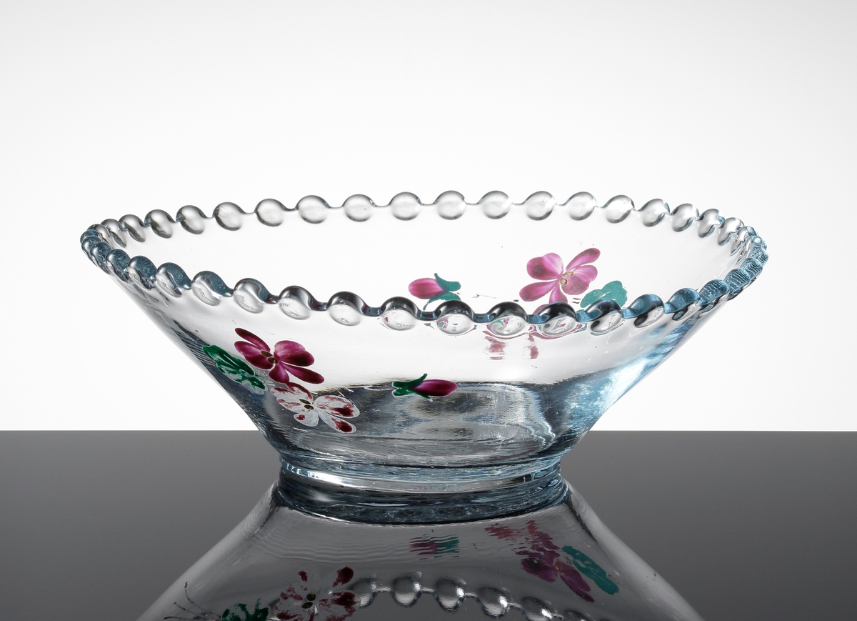 Rund pressglasskål med pärlkant, samt pärlor i relief i botten. Svagt blåtonat glas. Handmålad dekor bestående av två buketter med vinröda violer och gröna blad.