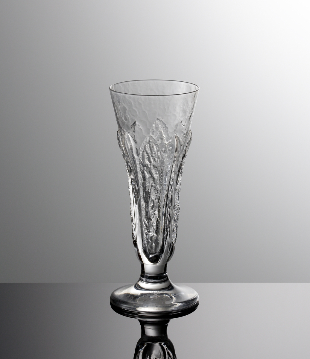 Snaps- sherry- eller likörglas ur servis "Eden". Formgiven av  Lars Hellsten. Art.nr. 8700-121 anges i katalogen "Nyheter 1975" som ett glas med tre användningsområden. Konisk kupe, något utsvängd mynning. Stående stiliserade blad runt skålen. Kort ben och relativt tjock fot.