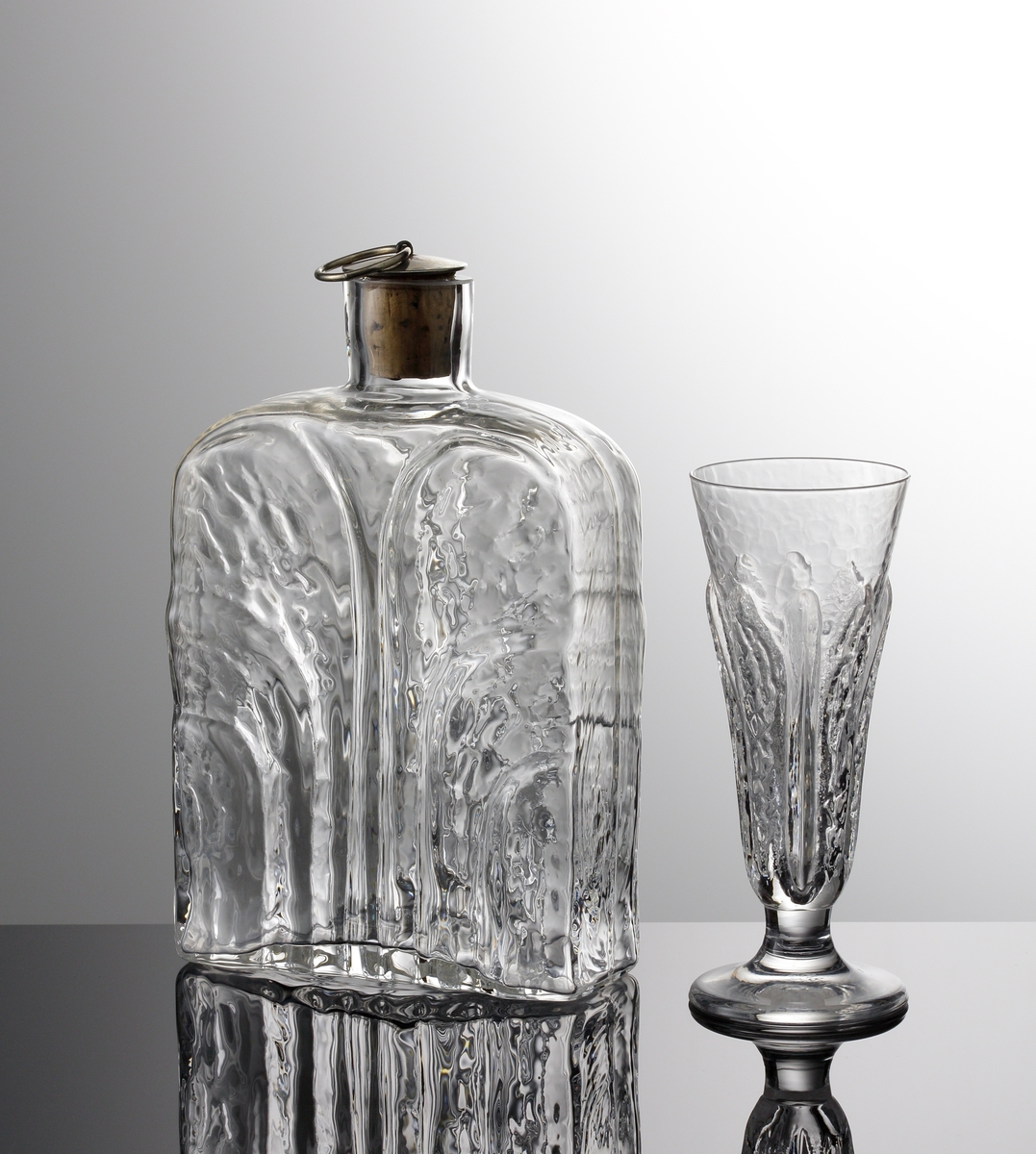 Snaps- sherry- eller likörglas ur servis "Eden". Formgiven av  Lars Hellsten. Art.nr. 8700-121 anges i katalogen "Nyheter 1975" som ett glas med tre användningsområden. Konisk kupe, något utsvängd mynning. Stående stiliserade blad runt skålen. Kort ben och relativt tjock fot.
