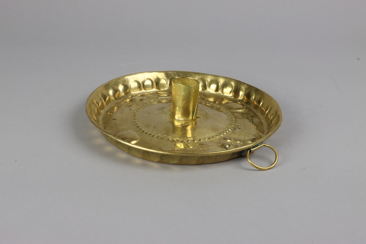 Ljusplåt av mässingsplåt, rund fatliknande form med något utvikt brätte. Cylindrisk ljuspipa av mässingsplåt, fastnitad i centrum av plåten med tre nitar. Driven och punsad dekor i form av klot, pärlbård och blomsterslingor. Från utsidan fastnitad ögla för upphängning.