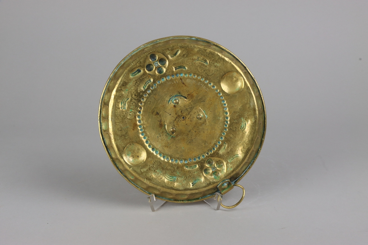 Ljusplåt av mässingsplåt, rund fatliknande form med något utvikt brätte. Cylindrisk ljuspipa av mässingsplåt, fastnitad i centrum av plåten med tre nitar. Driven och punsad dekor i form av klot, pärlbård och blomsterslingor. Från utsidan fastnitad ögla för upphängning.