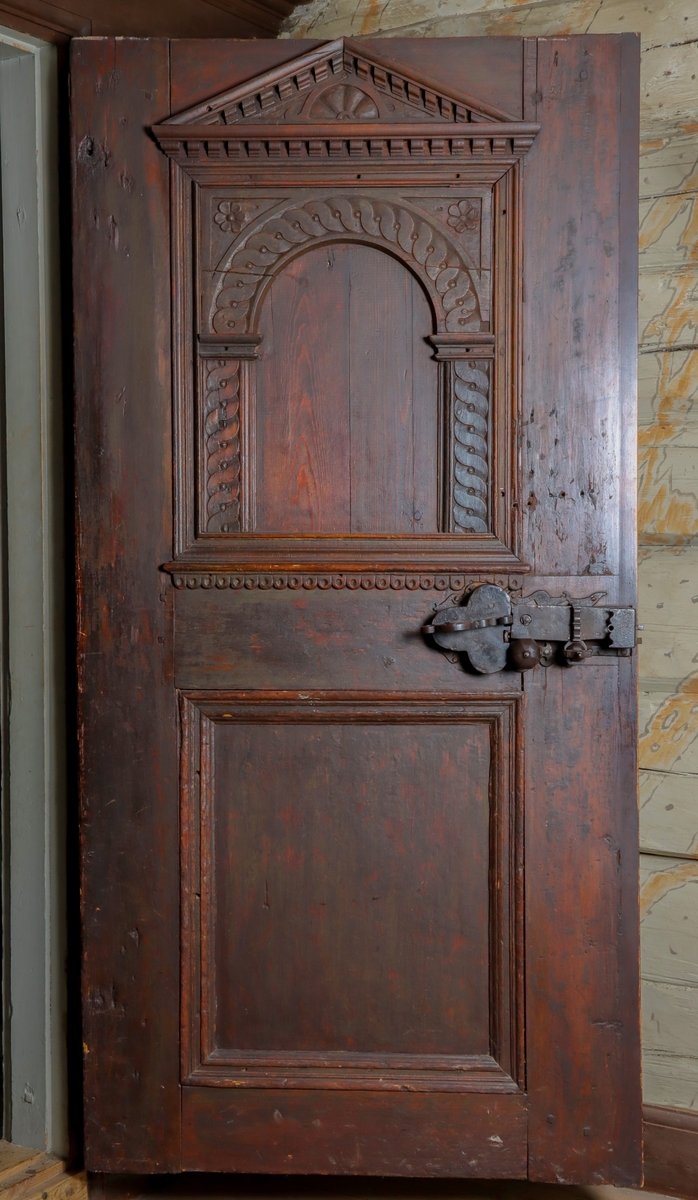 Helfransk ramverksdörr av trä, troligen fura, med två utanpåliggande speglar. Den övre spegelns fyllning med valvornament av skuret flätband samt skurna blommor i övre hörnen. Över valvbågen en triangulär fronton med gesims med skuret tandsnitt. Skuren rundbåge med blomma i frontonens mitt. Ornamenterat lås, trycke och nyckelskylt av järn. Hela dörren målad i en tunn rödbrun färg. Gångjärn av smide formade som dekorativa ornament.