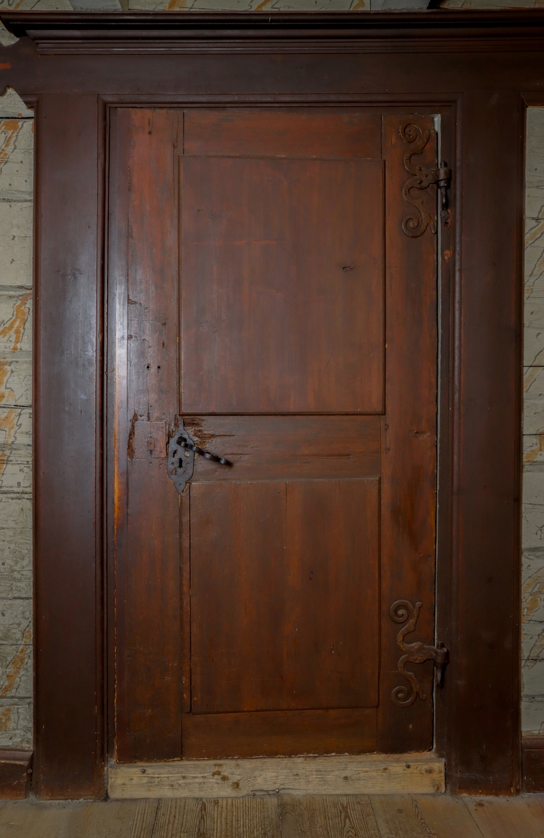 Helfransk ramverksdörr av trä, troligen fura, med två utanpåliggande speglar. Den övre spegelns fyllning med valvornament av skuret flätband samt skurna blommor i övre hörnen. Över valvbågen en triangulär fronton med gesims med skuret tandsnitt. Skuren rundbåge med blomma i frontonens mitt. Ornamenterat lås, trycke och nyckelskylt av järn. Hela dörren målad i en tunn rödbrun färg. Gångjärn av smide formade som dekorativa ornament.
