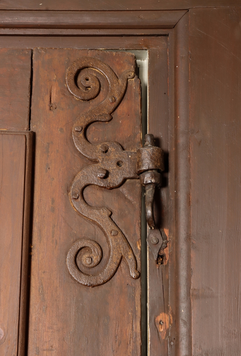 Helfransk ramverksdörr av trä, troligen fura, med två utanpåliggande speglar. Den övre spegelns fyllning med valvornament av skuret flätband samt skurna blommor i övre hörnen. Över valvbågen en triangulär fronton med gesims med skuret tandsnitt. Skuren rundbåge med blomma i frontonens mitt. Ornamenterat lås, trycke och nyckelskylt av järn. Hela dörren målad i en tunn rödbrun färg. Gångjärn av smide formade som dekorativa ornament.