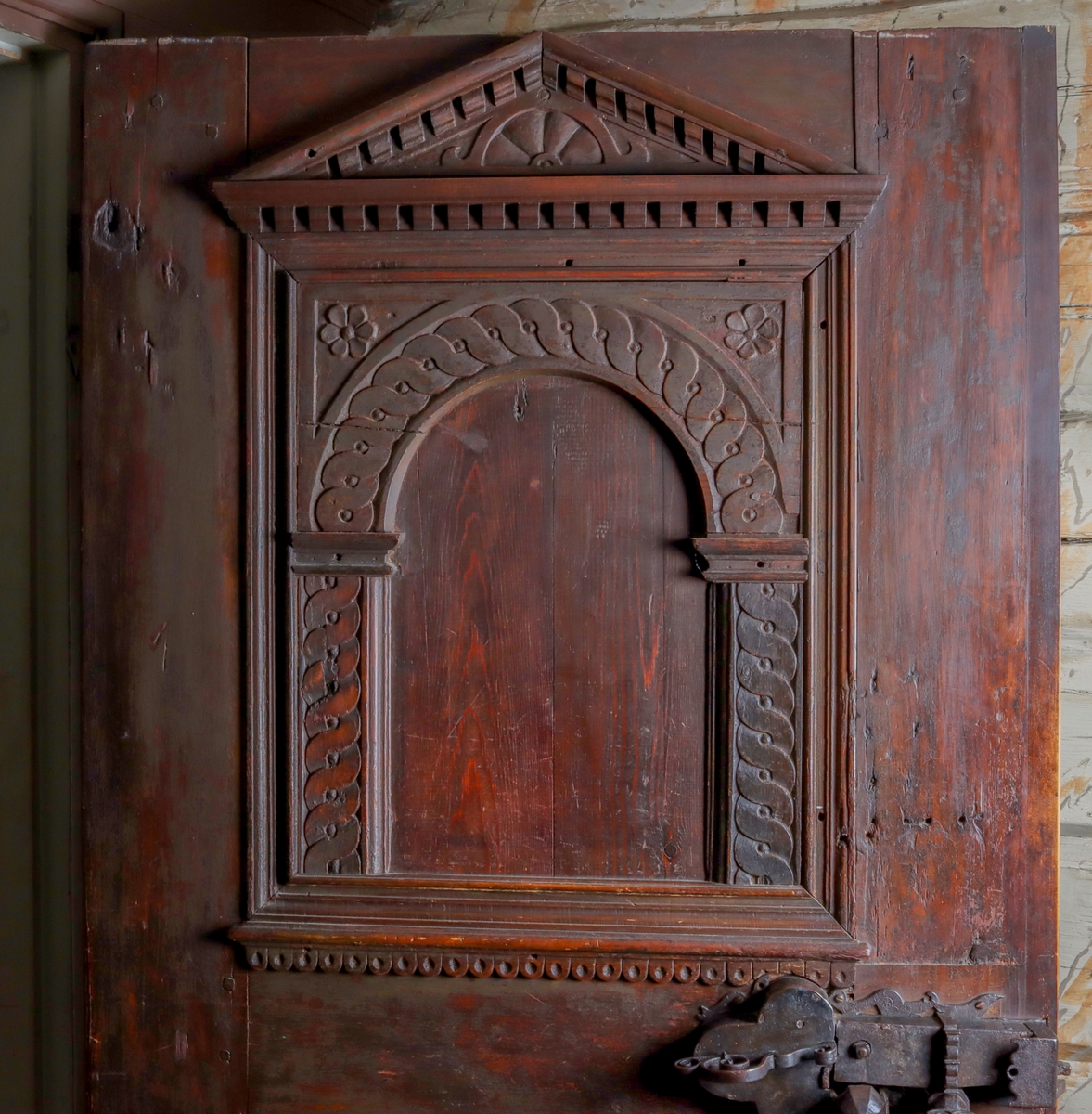 Helfransk ramverksdörr av trä, troligen fura, med två utanpåliggande speglar. Den övre spegelns fyllning med valvornament av skuret flätband samt skurna blommor i övre hörnen. Över valvbågen en triangulär fronton med gesims med skuret tandsnitt. Skuren rundbåge med blomma i frontonens mitt. Ornamenterat lås, trycke och nyckelskylt av järn. Hela dörren målad i en tunn rödbrun färg. Gångjärn av smide formade som dekorativa ornament.