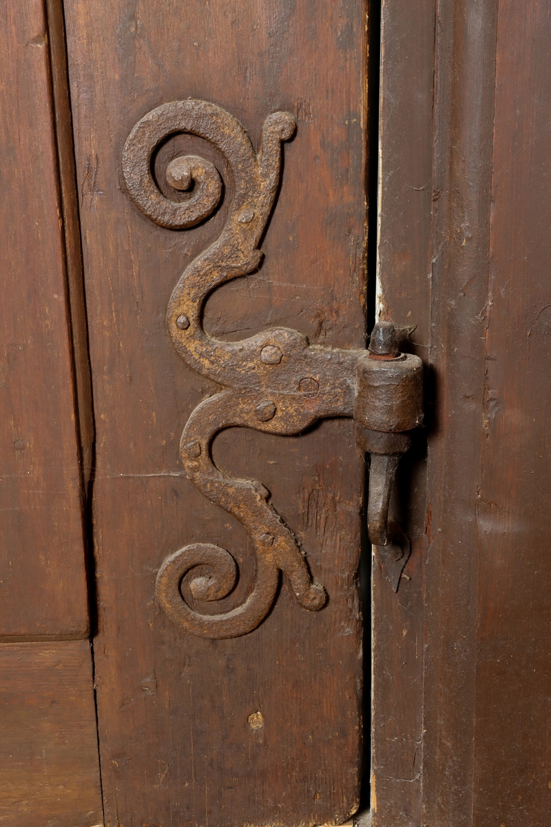 Helfransk ramverksdörr av trä, troligen fura, med två utanpåliggande speglar. Den övre spegelns fyllning med valvornament av skuret flätband samt skurna blommor i övre hörnen. Över valvbågen en triangulär fronton med gesims med skuret tandsnitt. Skuren rundbåge med blomma i frontonens mitt. Ornamenterat lås, trycke och nyckelskylt av järn. Hela dörren målad i en tunn rödbrun färg. Gångjärn av smide formade som dekorativa ornament.