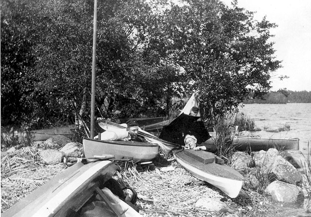 Fyra unga män vilar i kanoter som dragits upp på land. "Läger på Fredagsholmen 1906".