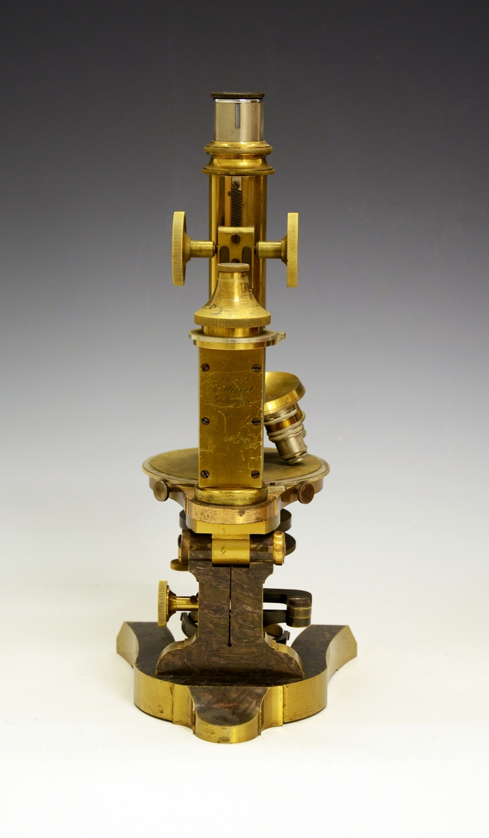 Mikroskop i kasse. Fra protokollen: Mikroskop. Stativet av massivt metall med hesteskoformet fot. Øverst på stativet røret (tubus) med objektiv og okular. Røret reguleres opp og ned ved drev og tannstang (grov innstilling). Bak røret skruehode for fin-innstilling. Mikroskopet har revolver med 4 forskjellige objektiver. Under disse bordet, som er en dreibar sirkulær skive med hull i midten hvorigjennom det sendes lys gjennom objektet fra et belysningsapparat under bordet. Nederst et dreielig plan- og hulspeil, over dette er blender som reguleres med tannstang og drev. Over blenderen kondensor med linser. På ryggen av stativet er inngravert: "No 1856 R. Winkel Göttingen 1891". Mikroskopet er plassert i et treskap med sidehengslet dør som har lås (nøkkelen mangler) og metallhandtak på toppstykket. Inni skapet og på innsiden av døren profilerte treknotter foret med filt, som holder mikrioskopet på plass. Profilerte lister (skinner) bærer et penalhus av tre med skyvelokk, en foret og trukket tre-eske som har et toppstykke med 5 hull (Esken er merket "E. Leitz, Wetzlar") og øverst en treplate med 3 større og 2 mindre hull. På toppen av døren mrk. "132986". Løst inni skapet en kondensor og en metallhylse.