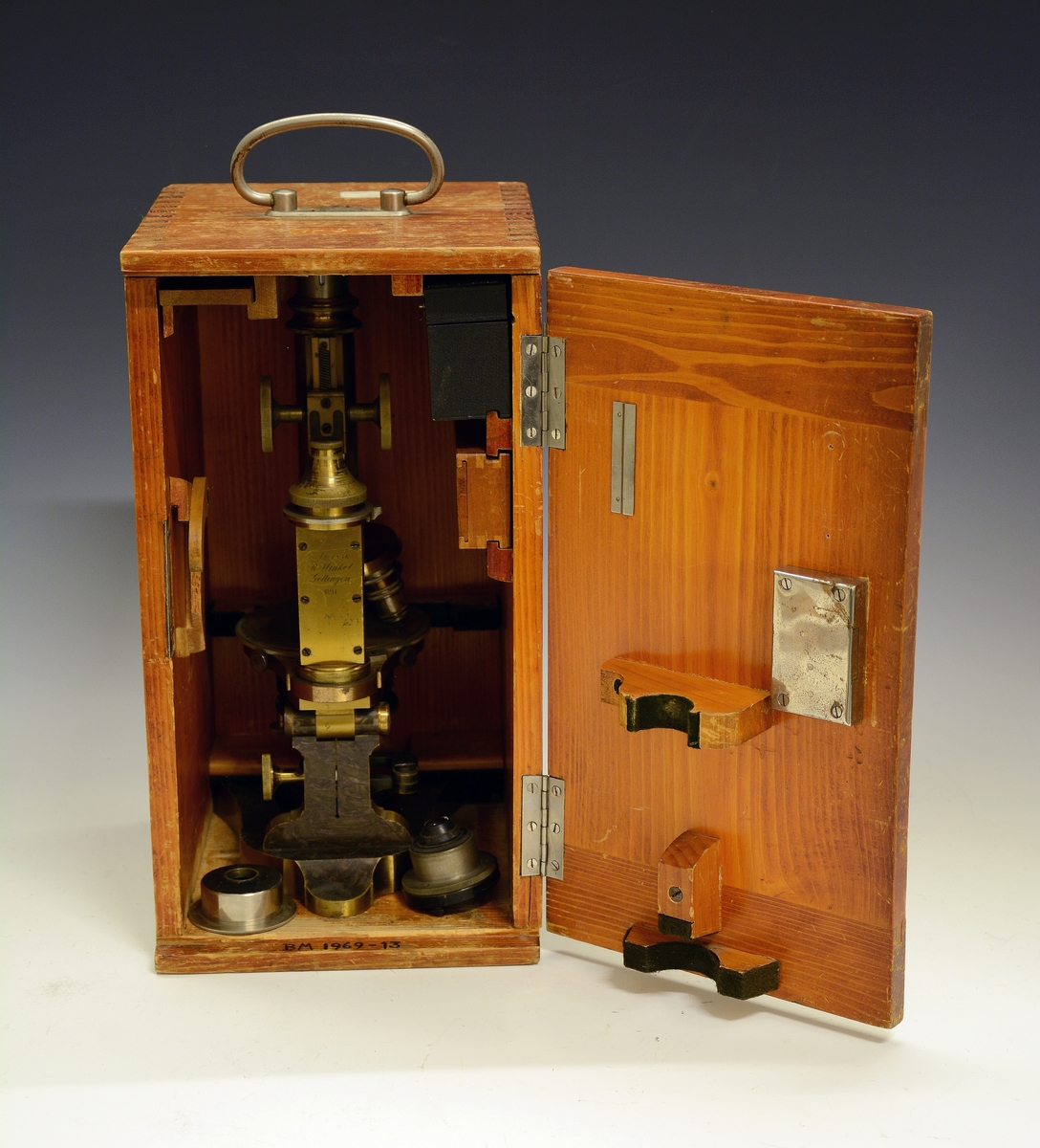 Mikroskop i kasse. Fra protokollen: Mikroskop. Stativet av massivt metall med hesteskoformet fot. Øverst på stativet røret (tubus) med objektiv og okular. Røret reguleres opp og ned ved drev og tannstang (grov innstilling). Bak røret skruehode for fin-innstilling. Mikroskopet har revolver med 4 forskjellige objektiver. Under disse bordet, som er en dreibar sirkulær skive med hull i midten hvorigjennom det sendes lys gjennom objektet fra et belysningsapparat under bordet. Nederst et dreielig plan- og hulspeil, over dette er blender som reguleres med tannstang og drev. Over blenderen kondensor med linser. På ryggen av stativet er inngravert: "No 1856 R. Winkel Göttingen 1891". Mikroskopet er plassert i et treskap med sidehengslet dør som har lås (nøkkelen mangler) og metallhandtak på toppstykket. Inni skapet og på innsiden av døren profilerte treknotter foret med filt, som holder mikrioskopet på plass. Profilerte lister (skinner) bærer et penalhus av tre med skyvelokk, en foret og trukket tre-eske som har et toppstykke med 5 hull (Esken er merket "E. Leitz, Wetzlar") og øverst en treplate med 3 større og 2 mindre hull. På toppen av døren mrk. "132986". Løst inni skapet en kondensor og en metallhylse.