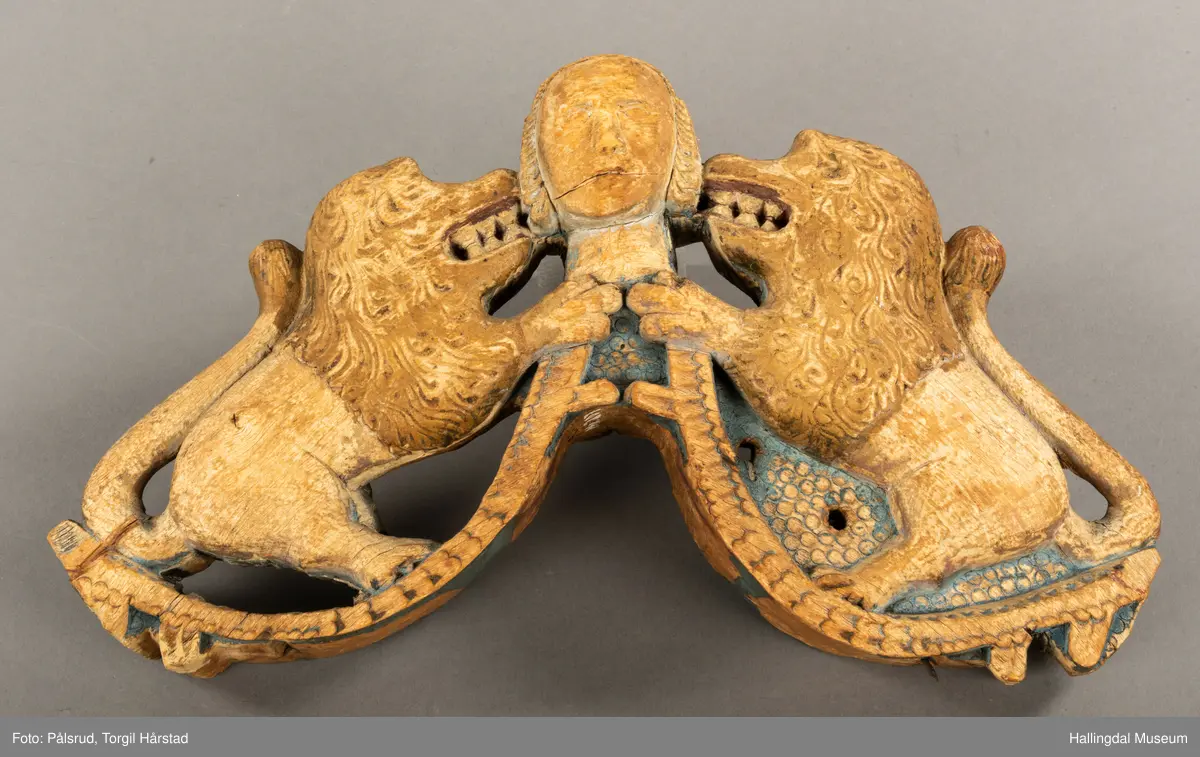 Tre, skåret i polykrom. Avslitt overmaling på en side, bedre bevart på baksiden. Toppfigur, mannshode; på hver side en løve med åpent gap.Halene ligger opp langs ryggene. Tømringene dannes ved halens begynnelse. Det kan virke som om endestykkene er brutt av, og høvren mangler bæreputer.