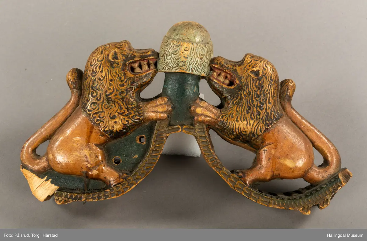 Tre, skåret i polykrom. Avslitt overmaling på en side, bedre bevart på baksiden. Toppfigur, mannshode; på hver side en løve med åpent gap.Halene ligger opp langs ryggene. Tømringene dannes ved halens begynnelse. Det kan virke som om endestykkene er brutt av, og høvren mangler bæreputer.