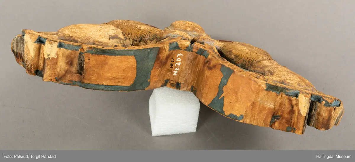 Tre, skåret i polykrom. Avslitt overmaling på en side, bedre bevart på baksiden. Toppfigur, mannshode; på hver side en løve med åpent gap.Halene ligger opp langs ryggene. Tømringene dannes ved halens begynnelse. Det kan virke som om endestykkene er brutt av, og høvren mangler bæreputer.