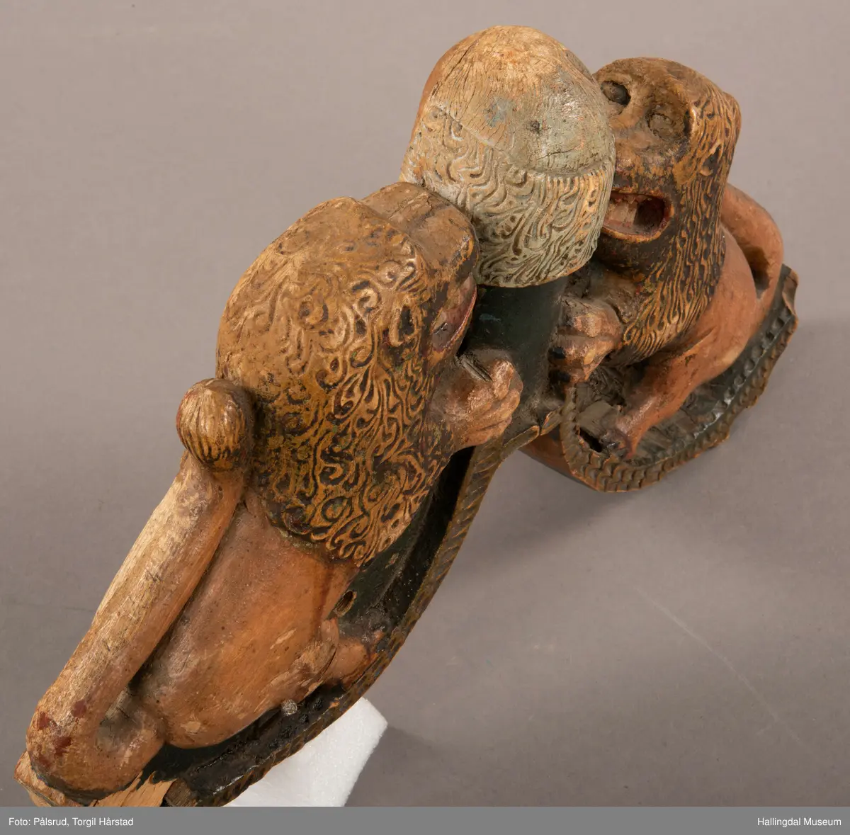 Tre, skåret i polykrom. Avslitt overmaling på en side, bedre bevart på baksiden. Toppfigur, mannshode; på hver side en løve med åpent gap.Halene ligger opp langs ryggene. Tømringene dannes ved halens begynnelse. Det kan virke som om endestykkene er brutt av, og høvren mangler bæreputer.