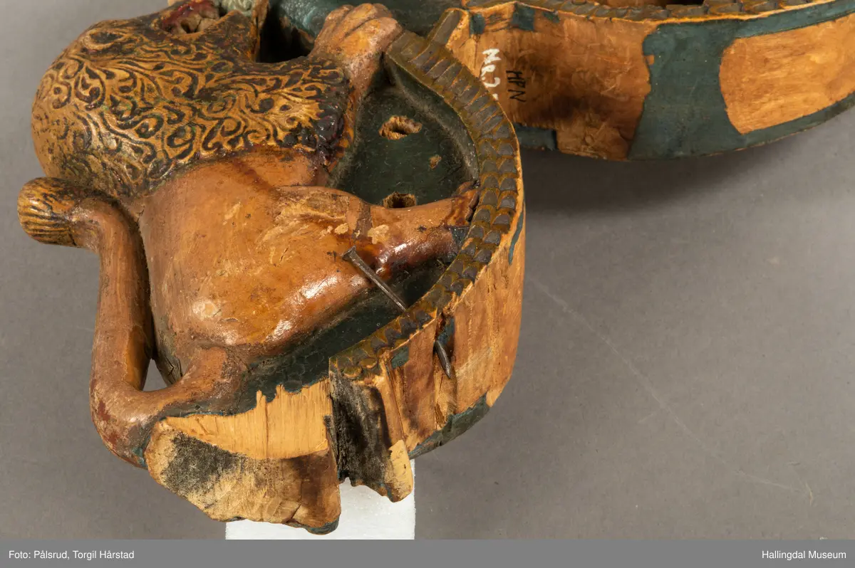 Tre, skåret i polykrom. Avslitt overmaling på en side, bedre bevart på baksiden. Toppfigur, mannshode; på hver side en løve med åpent gap.Halene ligger opp langs ryggene. Tømringene dannes ved halens begynnelse. Det kan virke som om endestykkene er brutt av, og høvren mangler bæreputer.