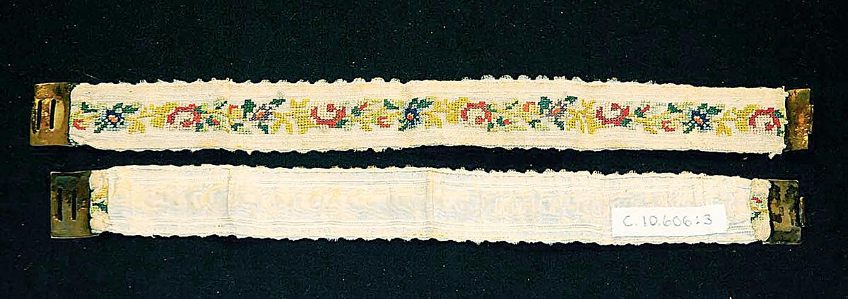 Ett par strumpeband med silkebroderi på fin stramalj. Motiv: Blommor och blad i rött, gult, rosa, lila, grönt mm. Baksida av vitt sidenband. Spännen av silver stämplade "AW"
Broderi på fin stramalj, gulvit med blommor och blad i rött, blått, grönt mm. Foder av vita sidenband. Spännen av silver, sign: A W. Längd: 28, br 2,7 cm. Föremålen (C10606:1 - 26) har tillhört donators svärmor, fru Agnes krokstedt, född Westöö, Slite
