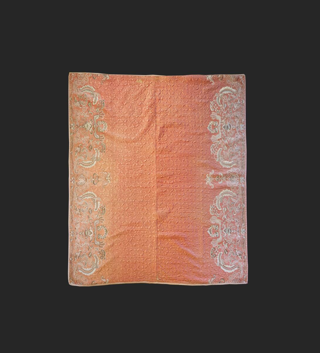 Av rosafärgad sidenatlas med foder av rosa bomullstyg, vadderat och stickat. Består av två broderade längder som ihopsytts. Stickningens mönster är fyra blad som bildar ett rutmönster över hela mittpartiet. Längs långsidorna en bård av flera nyanser i grått och vitt silke i schattérsöm, knutsöm, bottensömmar och stjälkstygn. L. 182 cm, B. 183 cm. Se även: C10594 och C11184 Denna typ av broderade sidentäcken från 1700-talets mitt och senare är ofta omsydda av s.k. "stubbar", vadderade kjolar, då dessa började komma ur bruk. Broderiet är mycket rikt och vittnar om stor skicklighet. Proveniens: Visbyköpmannen Jacob Lindström (1764-1818) hade i sina två äktenskap tre döttrar. När dessa gifte sig skänkte han dem förutom en präktig utstyrsel varsitt brudtäcke. Den andra dottern i ordningen, Anna Margaretha (1807-1891), gift med köpmannen och lantbrukaren Axel Hägg på Katthamra i Östergarn, fick detta täcke. Täcket har efter Anna Margaretha tillhört: 2) Dottern Maria Elisabeth Hägg (1837-1917), Katthamra. 3) Dottern Axelina Charlotta Jacobina (1843-1931), gift Sundell. 4) Axelinas dotter, Ester Kristina (f. 1882), gift Söderman, Katthammarsvik. År 1969 förvärvades det, genom flera sällskaps och fonders donationer, till Fornsalen. Täcket var under lång tid utställt på Fornsalen i utställningen "Gotland för 100 år sedan". Det visades i en dräktmonter tillsammans med de två andra brudtäckena från Jacob Lindströms döttrar. Läs mer: Ulla Melin, "Textila Släktklenoder: Några Anteckningar från Gotlands Hemslöjdsförenings Textilinventering 1944-1948", sid. 88-105, Gotländskt Arkiv 1948-49 & "Historien om tre brudtäcken", Utställningstext, Gotlands Museum
AV SIDEN, FODER AV ROSA BOMULLSTYG. VADDERAT OCH STICKAT. BESTÅR AV 2 BRODERADE LÄNGDER SOM HOPSYTTS. STICKNINGENS MÖNSTER ÄR 4 BLAD, SOM BILDAR ETT RUTMÖNSTER ÖVER HELA MITTPARTIET. LÄNGS LÅNGSIDORNA BÅRD AV FLERA NYANSER I GRÅTEIGT SILKE I SCHATTÉRSÖM, KNUTSÖM, BOTTENSÖMMAR OCH STJÄLKSTYGN. L. 182, BR. 183 CM. INKÖPT FRÅN FRU ESTER SÖDERMAN, KATTHAMMARSVIK,SE GOTL. ARKIV, 1948-49, SID 88.