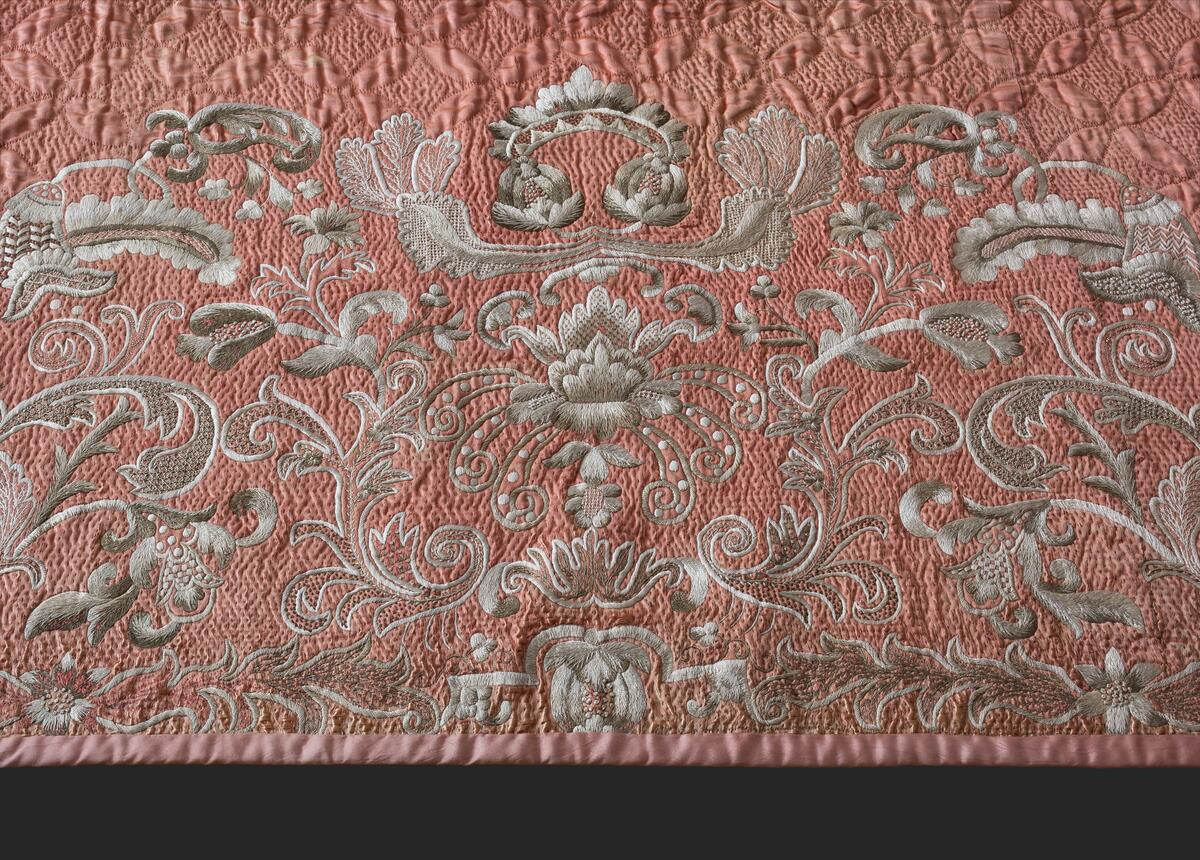 Av rosafärgad sidenatlas med foder av rosa bomullstyg, vadderat och stickat. Består av två broderade längder som ihopsytts. Stickningens mönster är fyra blad som bildar ett rutmönster över hela mittpartiet. Längs långsidorna en bård av flera nyanser i grått och vitt silke i schattérsöm, knutsöm, bottensömmar och stjälkstygn. L. 182 cm, B. 183 cm. Se även: C10594 och C11184 Denna typ av broderade sidentäcken från 1700-talets mitt och senare är ofta omsydda av s.k. "stubbar", vadderade kjolar, då dessa började komma ur bruk. Broderiet är mycket rikt och vittnar om stor skicklighet. Proveniens: Visbyköpmannen Jacob Lindström (1764-1818) hade i sina två äktenskap tre döttrar. När dessa gifte sig skänkte han dem förutom en präktig utstyrsel varsitt brudtäcke. Den andra dottern i ordningen, Anna Margaretha (1807-1891), gift med köpmannen och lantbrukaren Axel Hägg på Katthamra i Östergarn, fick detta täcke. Täcket har efter Anna Margaretha tillhört: 2) Dottern Maria Elisabeth Hägg (1837-1917), Katthamra. 3) Dottern Axelina Charlotta Jacobina (1843-1931), gift Sundell. 4) Axelinas dotter, Ester Kristina (f. 1882), gift Söderman, Katthammarsvik. År 1969 förvärvades det, genom flera sällskaps och fonders donationer, till Fornsalen. Täcket var under lång tid utställt på Fornsalen i utställningen "Gotland för 100 år sedan". Det visades i en dräktmonter tillsammans med de två andra brudtäckena från Jacob Lindströms döttrar. Läs mer: Ulla Melin, "Textila Släktklenoder: Några Anteckningar från Gotlands Hemslöjdsförenings Textilinventering 1944-1948", sid. 88-105, Gotländskt Arkiv 1948-49 & "Historien om tre brudtäcken", Utställningstext, Gotlands Museum
AV SIDEN, FODER AV ROSA BOMULLSTYG. VADDERAT OCH STICKAT. BESTÅR AV 2 BRODERADE LÄNGDER SOM HOPSYTTS. STICKNINGENS MÖNSTER ÄR 4 BLAD, SOM BILDAR ETT RUTMÖNSTER ÖVER HELA MITTPARTIET. LÄNGS LÅNGSIDORNA BÅRD AV FLERA NYANSER I GRÅTEIGT SILKE I SCHATTÉRSÖM, KNUTSÖM, BOTTENSÖMMAR OCH STJÄLKSTYGN. L. 182, BR. 183 CM. INKÖPT FRÅN FRU ESTER SÖDERMAN, KATTHAMMARSVIK,SE GOTL. ARKIV, 1948-49, SID 88.