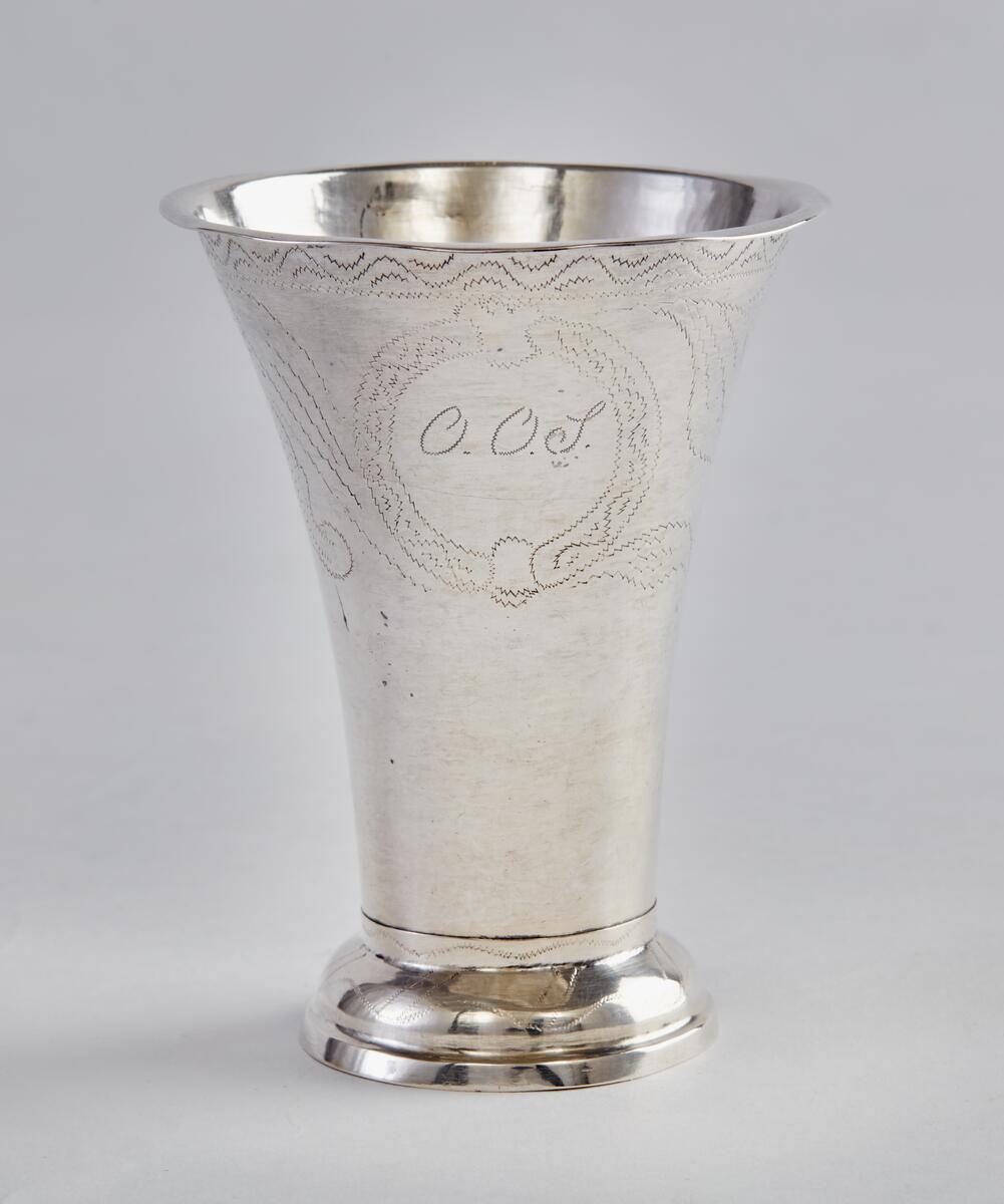Bägare, silver, med utsvängd, profilerad fot, vidgad uppåt med utvikt kant längst upp. Bägarens övre del försedd med dekor av vinkelböjd ranka, därunder bred blomranka med bl.a voluter och "OOS" inom blomkrans. Även den utsvängda foten dekorerad. På bägarens botten stämplar: "FUE", "A4", trekronorsstämpel, Falustämpel. FUE=Fredrik Ulrik Ekholm. A4=1831.

Från Västbergs by, Rättvik, se bilaga.