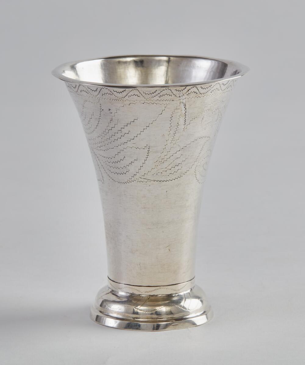 Bägare, silver, med utsvängd, profilerad fot, vidgad uppåt med utvikt kant längst upp. Bägarens övre del försedd med dekor av vinkelböjd ranka, därunder bred blomranka med bl.a voluter och "OOS" inom blomkrans. Även den utsvängda foten dekorerad. På bägarens botten stämplar: "FUE", "A4", trekronorsstämpel, Falustämpel. FUE=Fredrik Ulrik Ekholm. A4=1831.

Från Västbergs by, Rättvik, se bilaga.