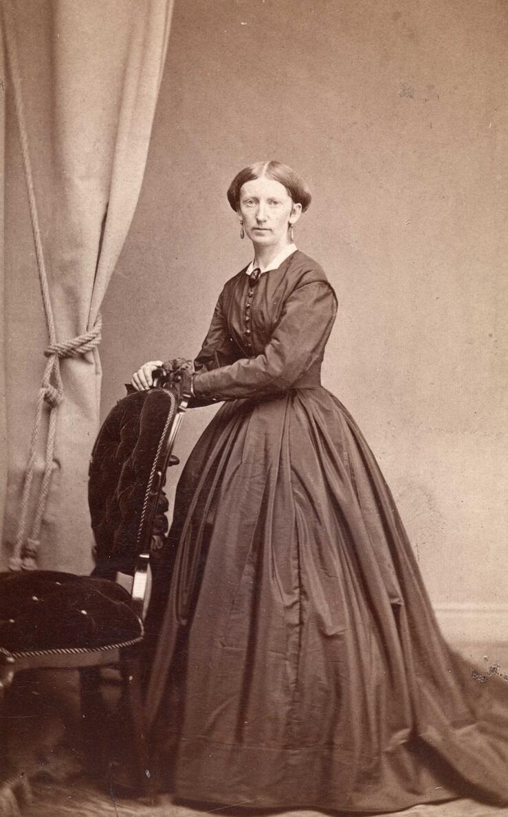 Fotografi av Thomasine Lie. Hun har på en flott kjole. I bakgrunnen ser vi en gardin til høyre. Hun holder hendene på baksiden av en stol.