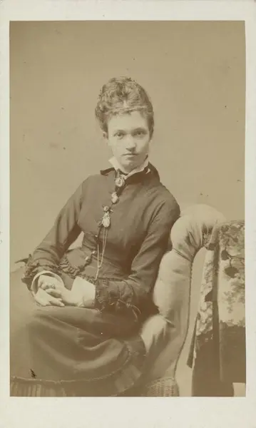 Fotografi av Jenny Sinclair. Hun sitter på en polstret stol og ser rett på kameraet.